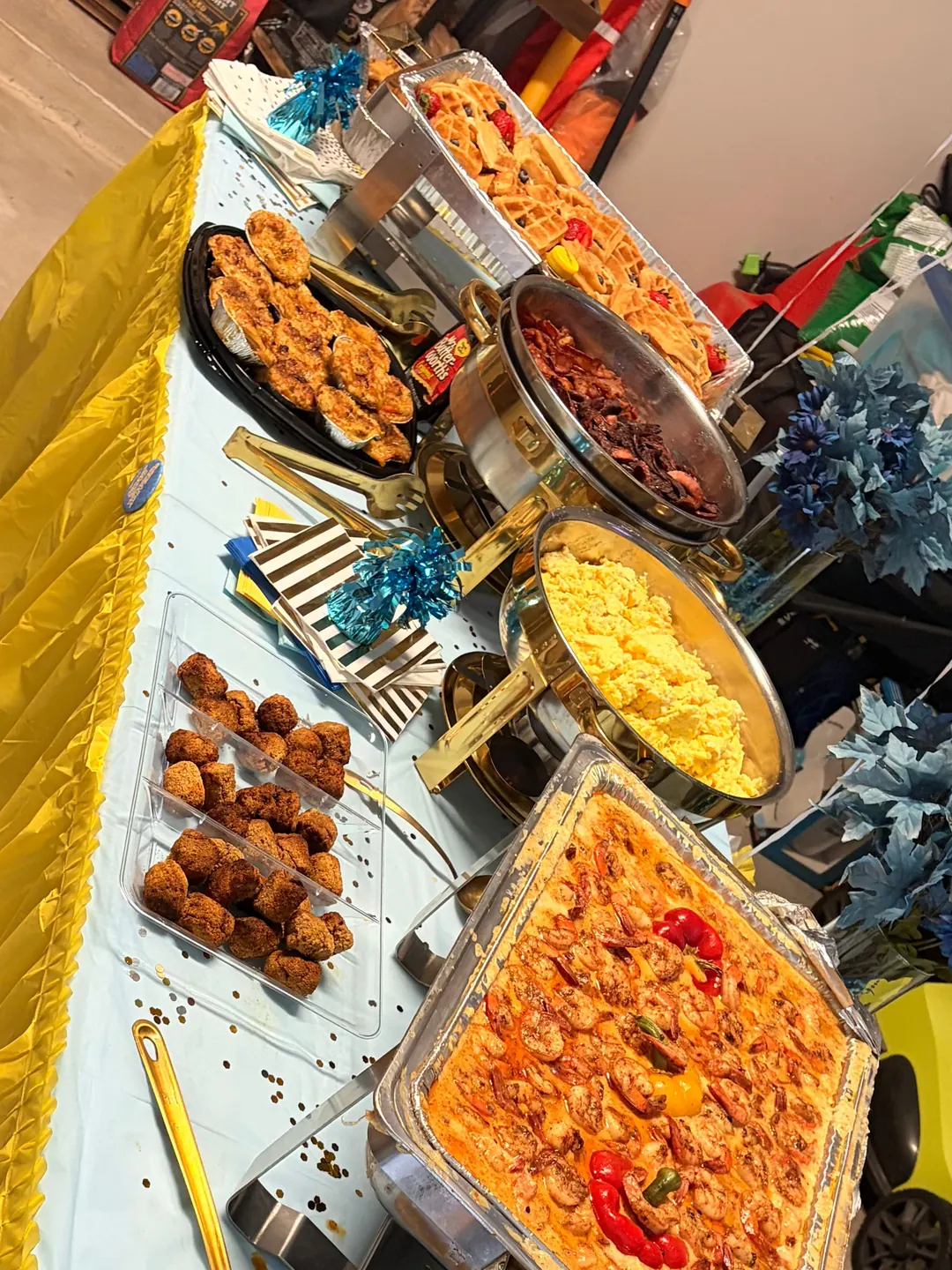 A brunch buffet with bacon, scrambled eggs, fried bites, waffles and Creole shrimp and grits on a blue and gold table